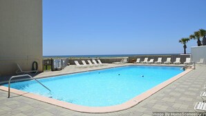 En rektangulär pool med kristallklart blått vatten, omgiven av solstolar och med utsikt över havet.