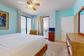 A hotel room with a bed, a ceiling fan, a TV, a wooden dresser, and a door leading to a balcony with a view of the ocean. - Gorgeous waterfront condo with pool, fitness room & WiFi (Navarre Beach)