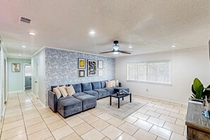 A spacious living room with a grey sectional sofa, a ceiling fan, and a large window with blinds. - Dog-friendly home steps to beach with 2 kitchens, dressing room & game room (Panama City Beach)