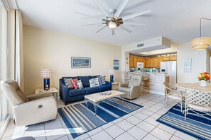 A living room with a sofa, recliner, coffee table, and dining area with chairs. There is a ceiling fan, kitchen in the background, and a window with curtains. - 7th-floor bright, open condo - close to dining with Gulf front views (Navarre Beach)