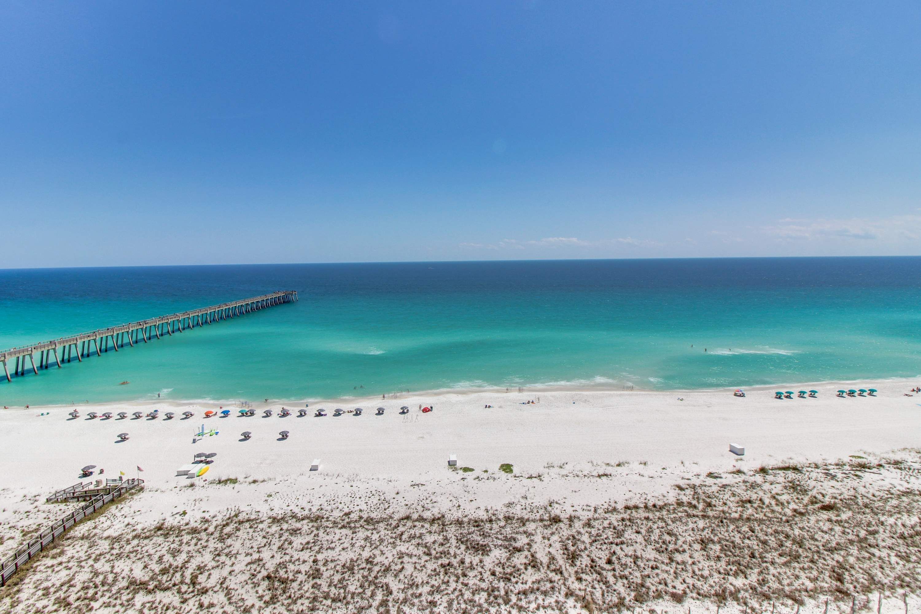 A beach with a pier extending into the ocean, a sandy beach with umbrellas and sun loungers, and a clear blue sky.