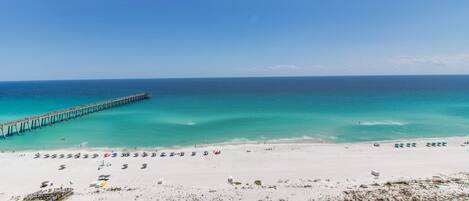 A beach with a pier extending into the ocean, a sandy beach with umbrellas and sun loungers, and a clear blue sky.