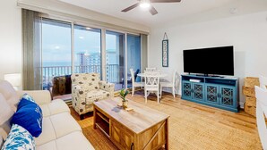 A spacious living room with a large window, a ceiling fan, a flat-screen TV, and a dining area. - Inviting beach view condo with balcony, amazing pool, gym, & W&D (Miramar Beach)