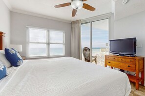 A hotel room with a large bed, a television on a wooden dresser, a ceiling fan, and a view of the ocean through a sliding glass door. - Picturesque Gulf-Front Condo Steps To Beach Gulfside Pool & Exercise Room (Fort Walton Beach)