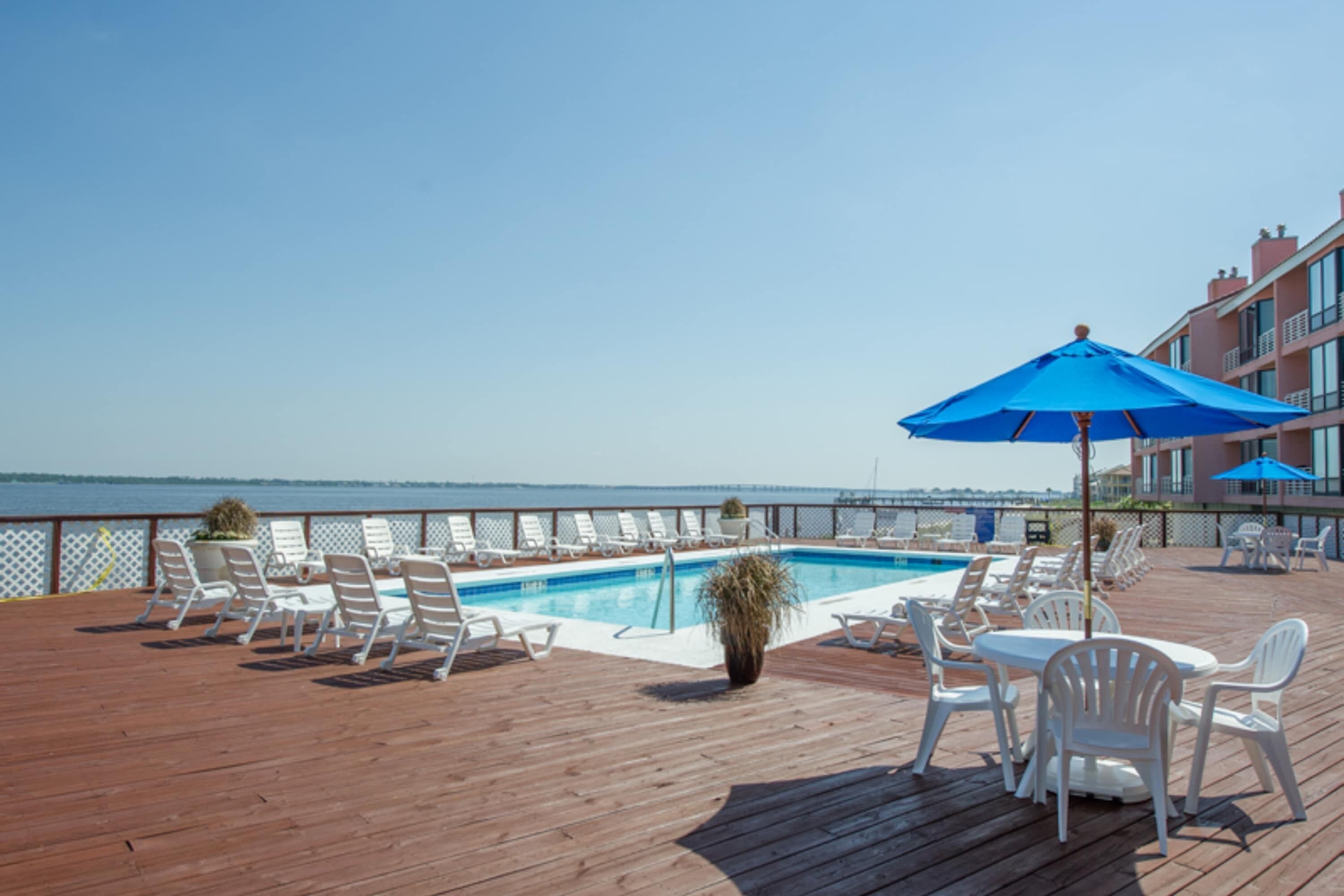A wooden deck with a pool, lounge chairs, and a table with a blue umbrella.