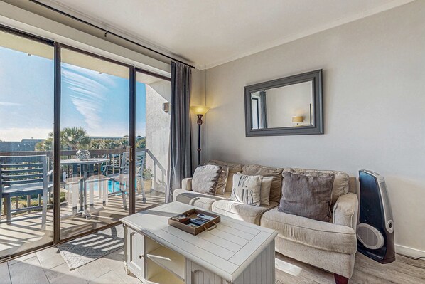 A living room with a sofa, a coffee table, a lamp, and a large window overlooking a balcony with outdoor furniture. - Cozy soundfront studio with shared pool, beach access & close to everything (Fort Walton Beach)