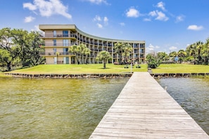 Un hotel con fachada curva, un muelle de madera que se adentra en una masa de agua y un área verde con árboles.