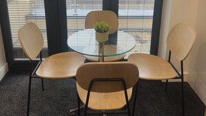 Huoneessa on lasipöytä, kaksi tuolia, ruukkukasvi ja suuret ikkunat, joissa on sälekaihtimet.