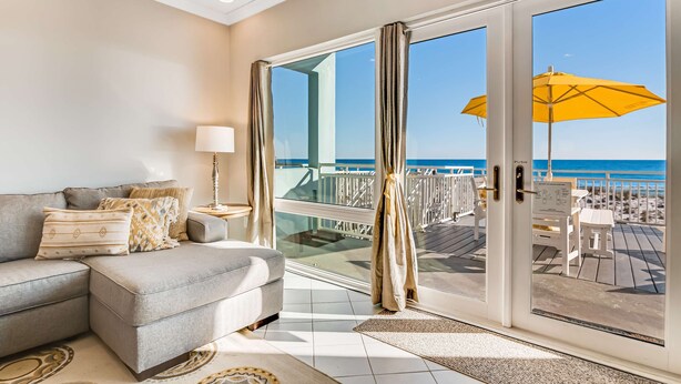 Ein modernes Wohnzimmer mit einem grauen Ecksofa, einem Beistelltisch mit einer Lampe und Blick auf den Strand durch große Glasfenster.