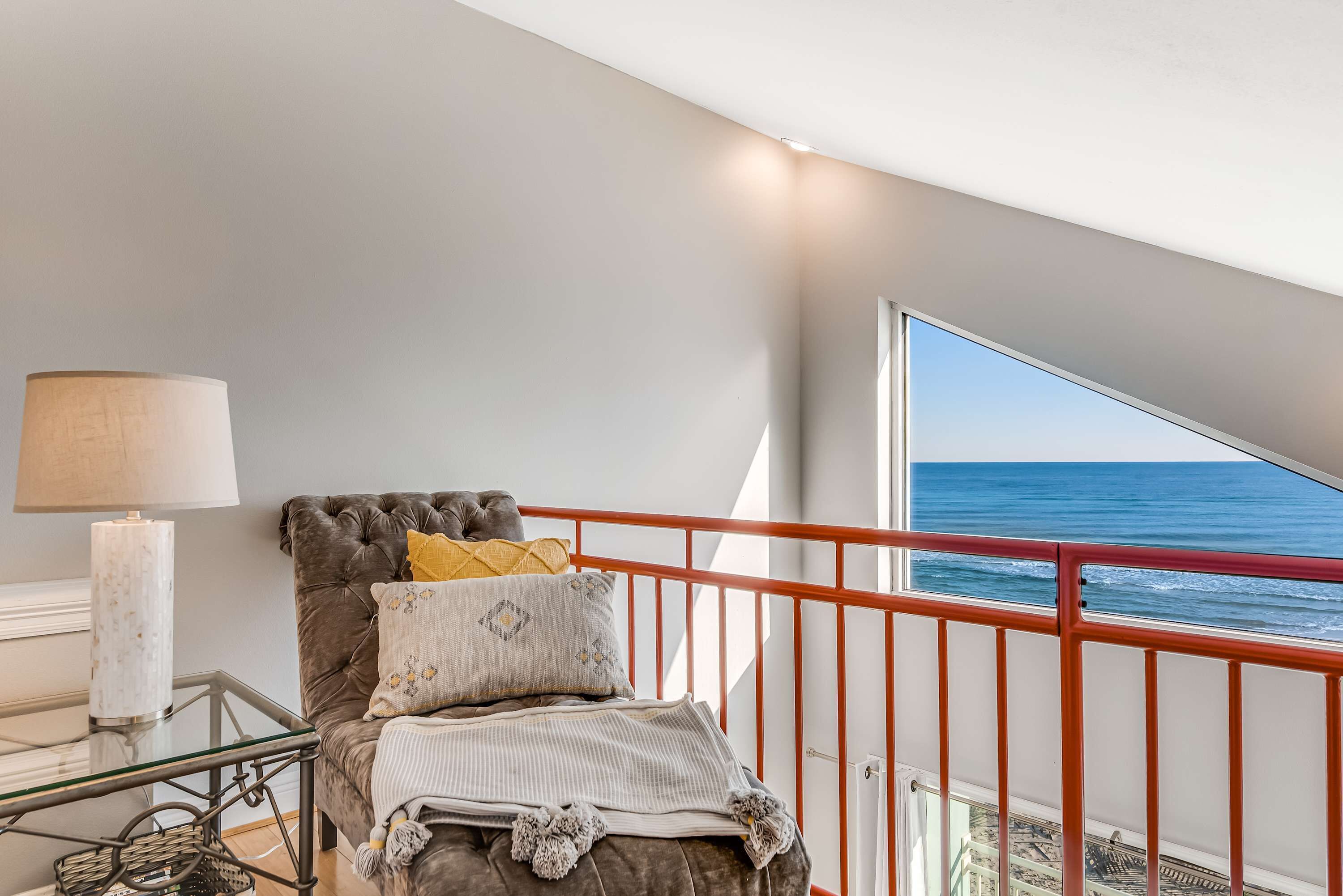 A balcony with a red railing, a bed with a striped blanket and decorative pillows, and a white lamp on a glass table.