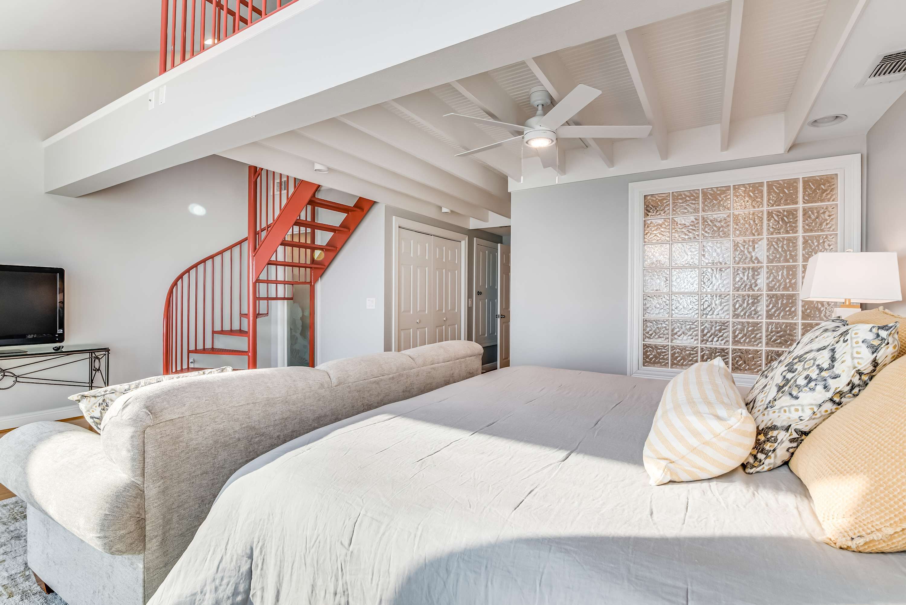 A bedroom with a large bed, a ceiling fan, a TV, a staircase, and a window with a decorative screen.