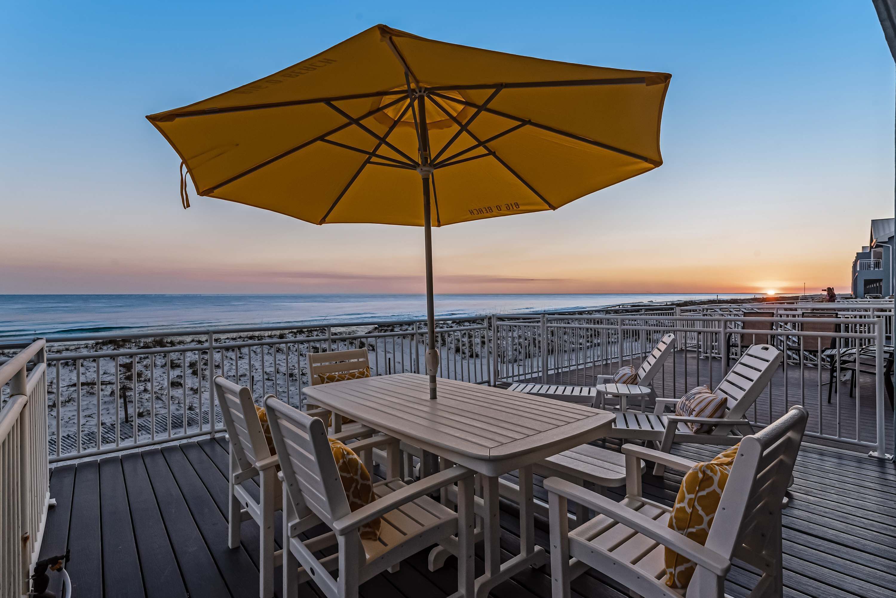 A deck with outdoor seating and a large yellow umbrella, overlooking the ocean at sunset.