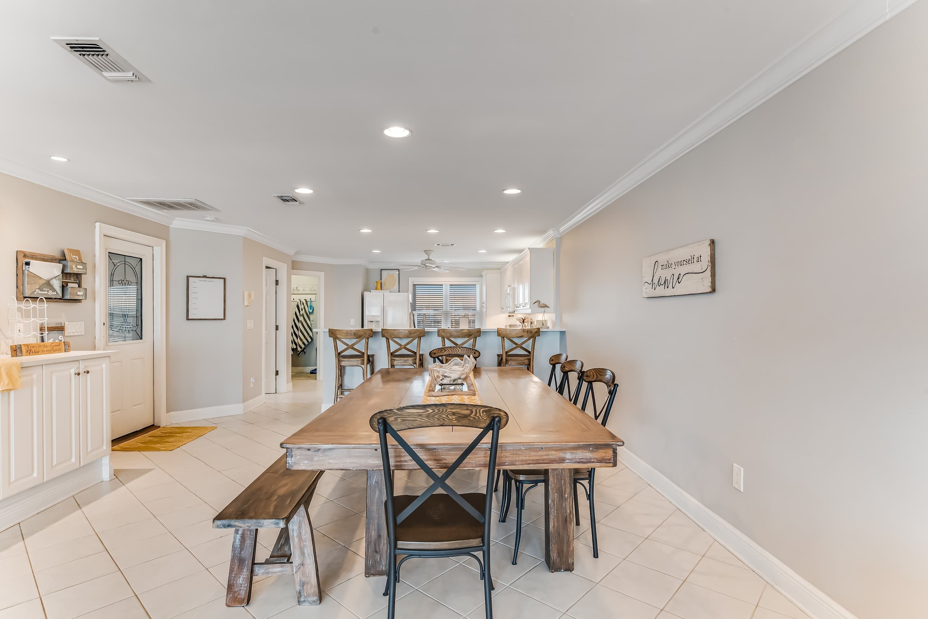 A spacious dining area with a long wooden table and multiple chairs, a kitchen in the background, and a sign on the wall.