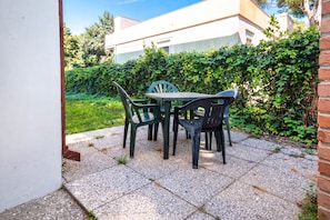 An outdoor patio with a table and chairs, surrounded by a hedge and a building in the background. - Cozy Flat With Outdoor Space (Rosolina)