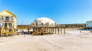 Una casa sulla spiaggia con struttura a cupola, terrazza in legno e una scala che conduce alla spiaggia.