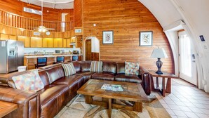 Un'ampia zona giorno con soffitto in legno curvo, un grande divano in pelle a forma di L, un tavolino da caffè in legno e una cucina sullo sfondo.