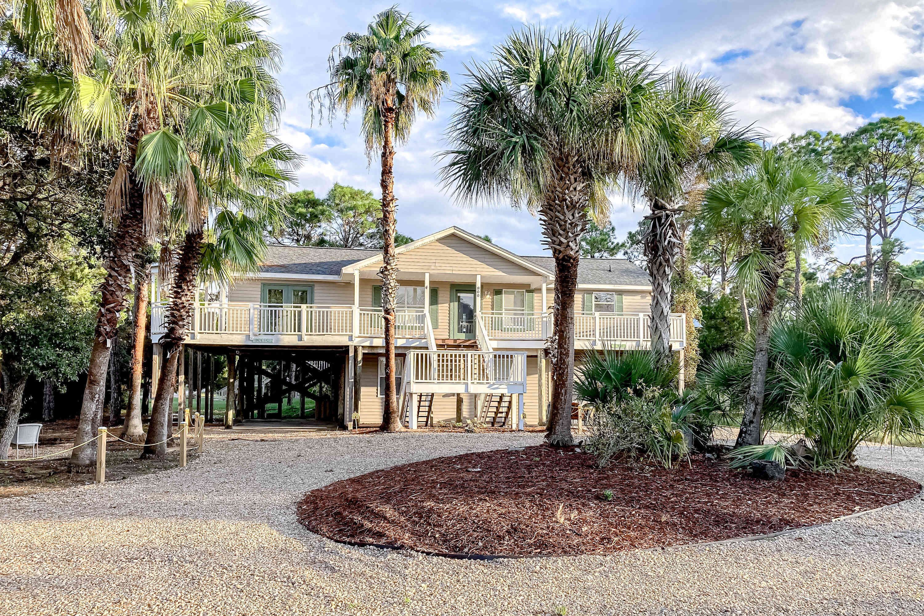 A two-story house with a covered porch, surrounded by palm trees and a gravel driveway.