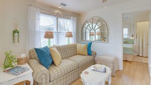 Un salon chaleureux avec un canapé à carreaux, une petite table blanche et une fenêtre ornée d’un vitrail décoratif.