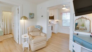 Une pièce comprenant un fauteuil beige, une petite table blanche, une commode bleue et un coin cuisine avec des placards blancs et un four à micro-ondes.