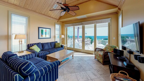A spacious living room with a large sofa, a coffee table, and a TV. There is a ceiling fan and a view of the ocean through large windows.