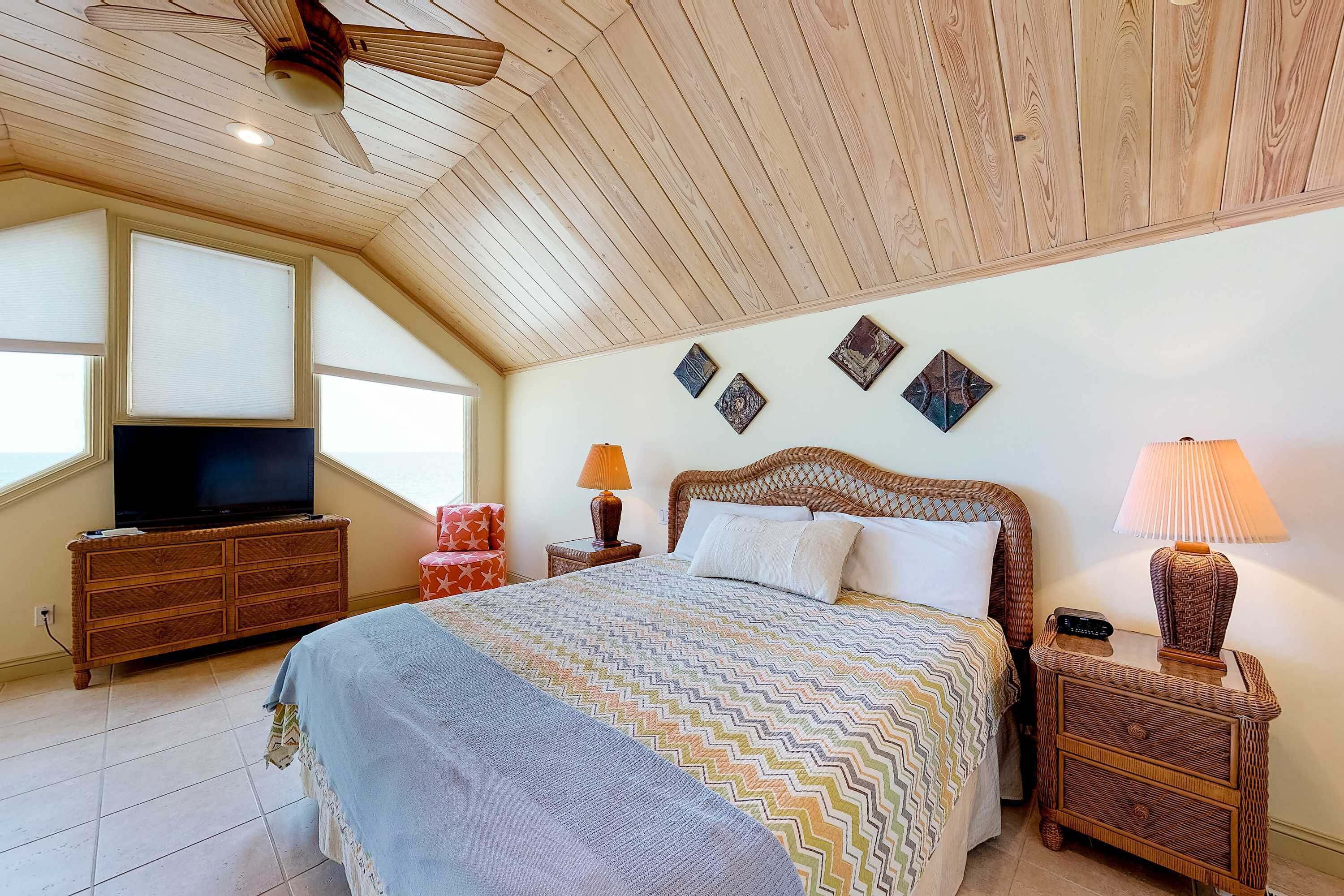 Une chambre avec un plafond en bois, un lit recouvert d’une couvre-lit à motifs, un téléviseur posé sur une commode en bois et une table de chevet avec une lampe.