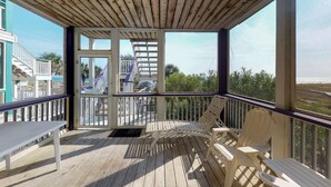 A wooden deck with a white table and two white chairs, overlooking a beach and a building with a blue exterior.