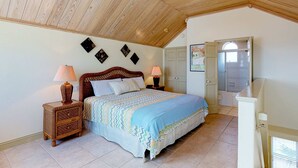 A bedroom with a wooden ceiling, a bed with a patterned bedspread, two bedside tables with lamps, and a door leading to a bathroom.