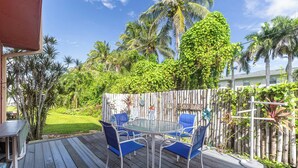 Een houten terras met een glazen tafel en blauwe stoelen, omringd door weelderige begroeiing en palmbomen.