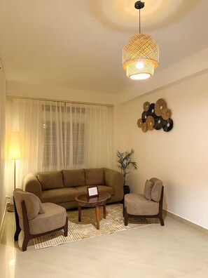 A living room with a beige sofa, two armchairs, a coffee table, a floor lamp, a wall-mounted decorative piece, and a hanging light fixture.
