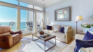 A spacious living room with a large window overlooking the sea, a ceiling fan, a sofa, a coffee table, and a side table with a lamp.