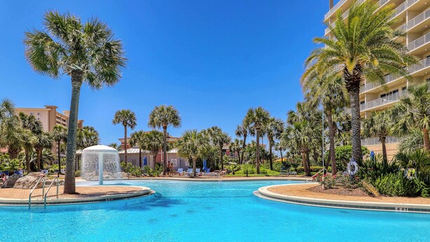 A swimming pool with palm trees and a water fountain, surrounded by a hotel building.
