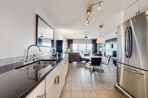 A modern kitchen with a stainless steel refrigerator, a black countertop, and a dining area with a table and chairs. - Sixth-Floor Oceanfront Condo with washer&dryer, Central AC, pool (Miramar Beach)