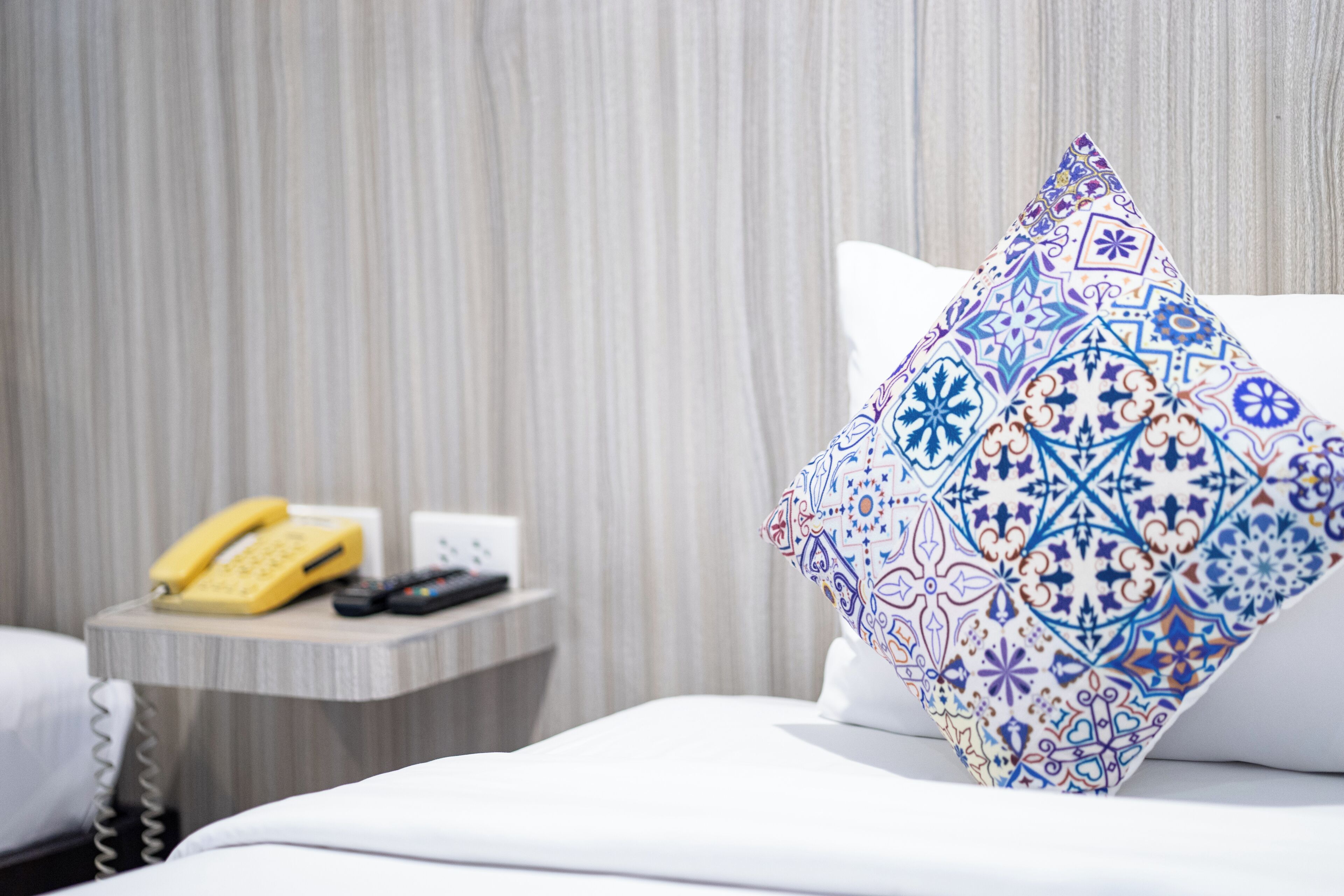 Quarto de hotel com uma cama, um travesseiro com padrão geométrico, mesa de cabeceira com telefone e controle remoto, e uma cortina ao fundo.