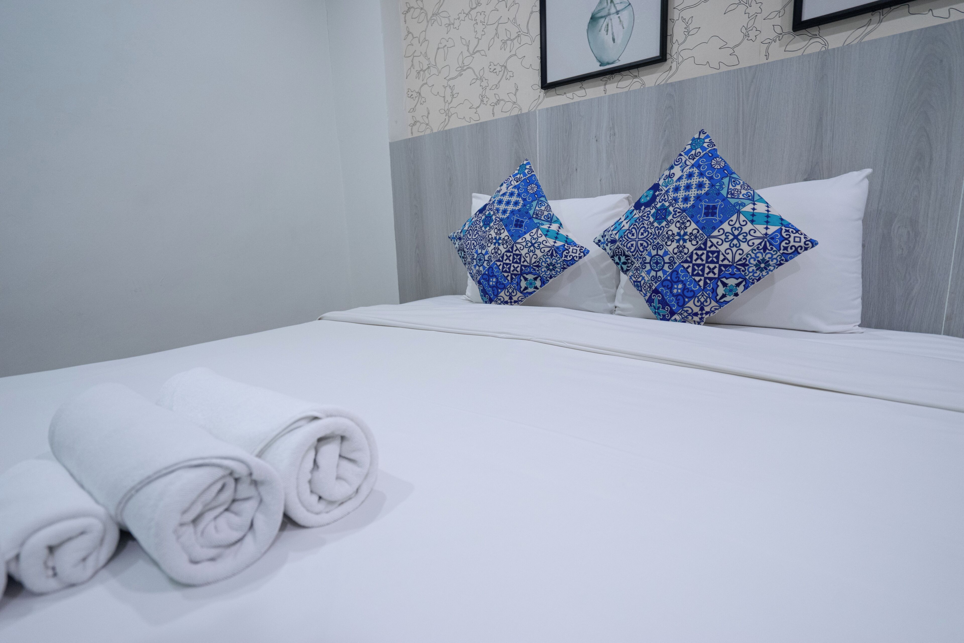 A neatly made bed with white linens and blue patterned pillows, a wooden headboard, and a framed picture on the wall.