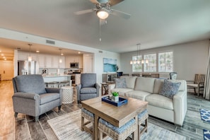 A spacious living area with a sofa, armchairs, a coffee table, and a dining table. There is a ceiling fan, kitchen cabinets, and a large window with blinds. - Blu 101 - Luxurious first-floor condo with hot tub, heated outdoor pool - near entertainment (Fort Walton Beach)