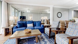 En stue med en blå sofa, et glasbord, fletstole og et stort spejl.