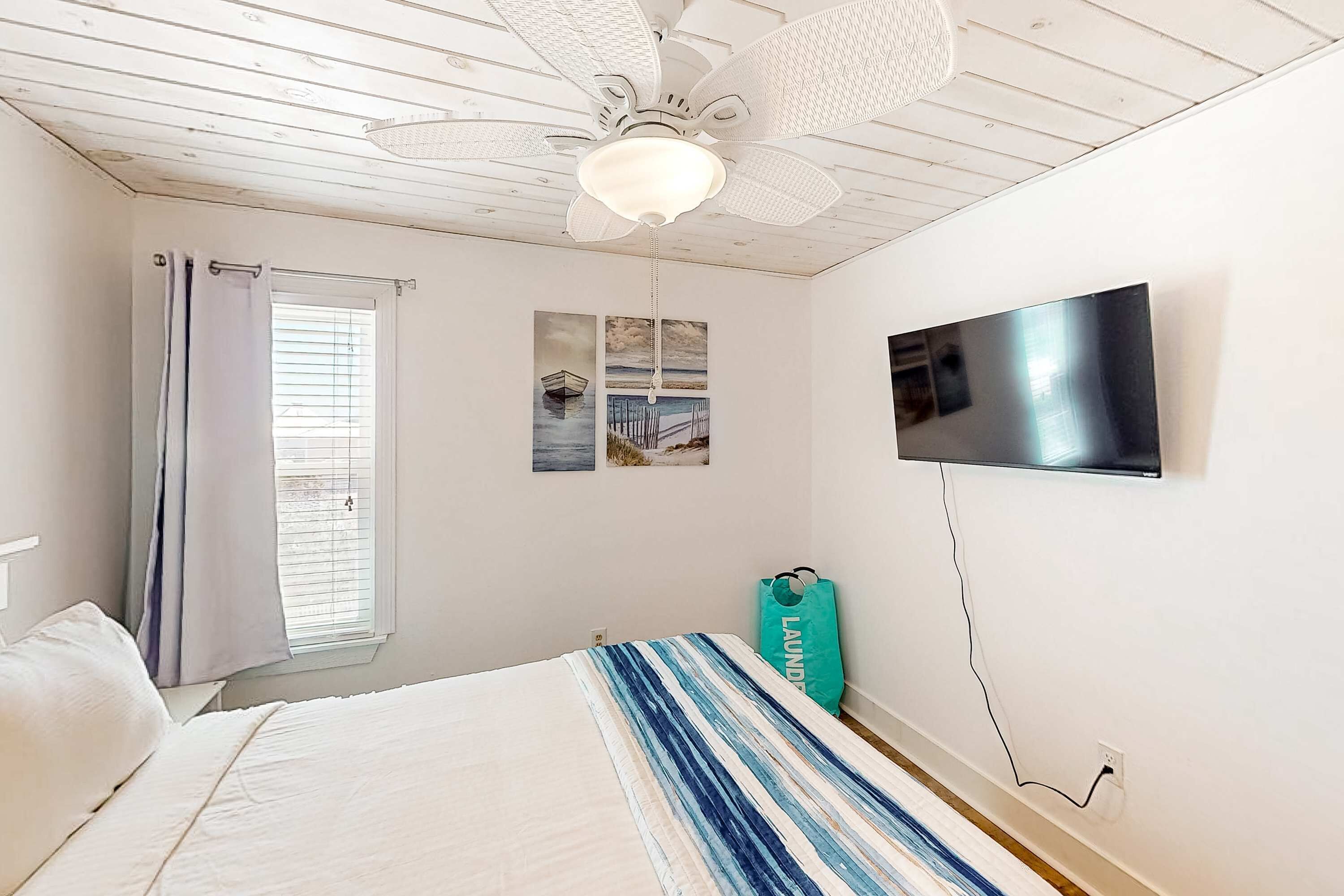 A bedroom with a bed, a ceiling fan, a TV mounted on the wall, and a window with curtains.