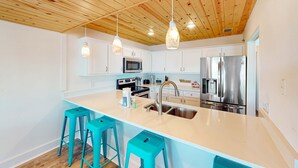 Una cocina moderna con techo de madera, armarios blancos, una isla central con taburetes de bar, electrodomésticos de acero inoxidable e iluminación colgante.