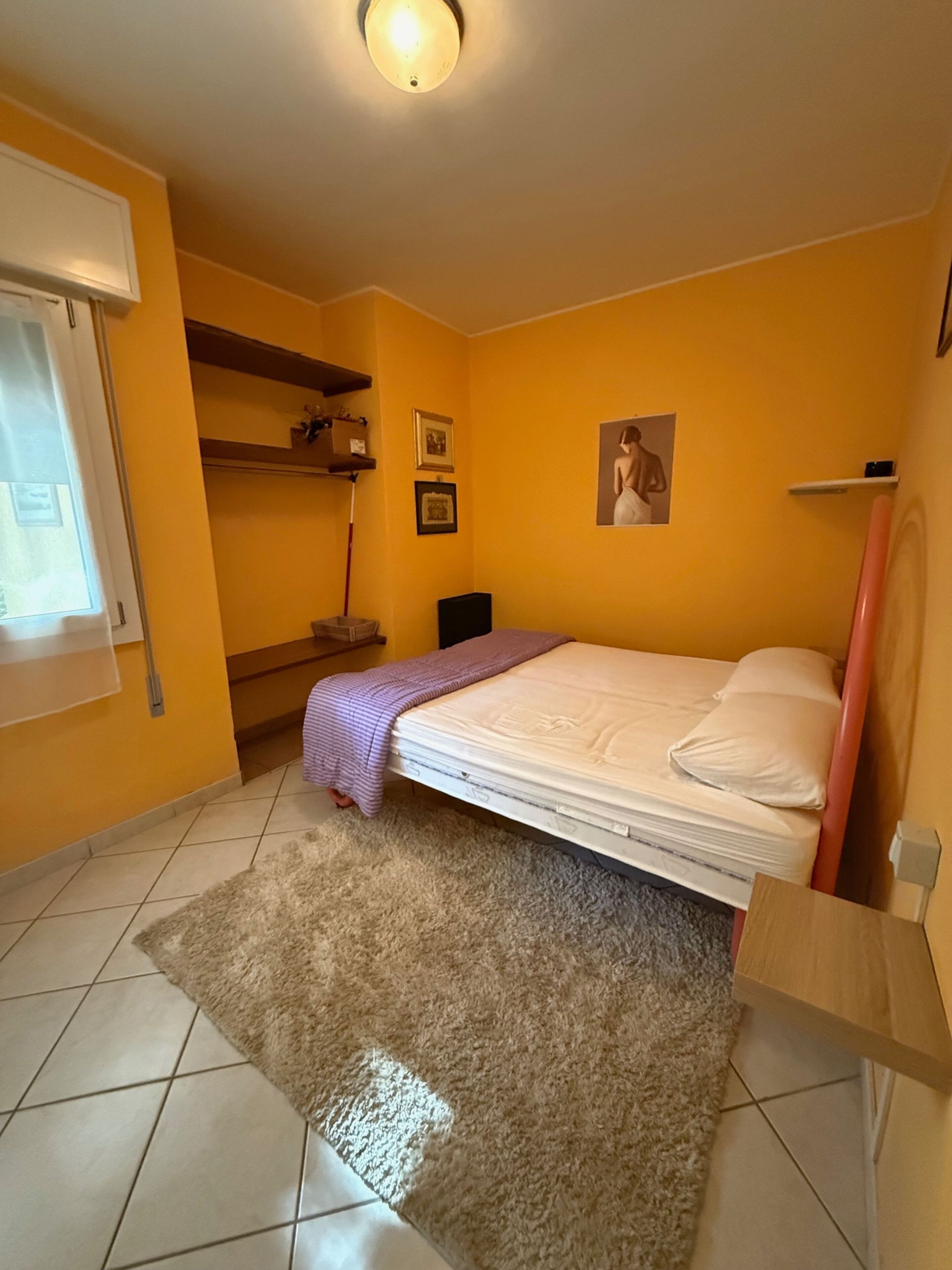 Une petite pièce lumineuse, avec un lit, une fenêtre et une étagère où sont posés des objets personnels.