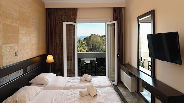 Ein Hotelzimmer mit zwei Betten, einem Fernseher, einem Balkon mit Tisch und Stühlen sowie Blick ins Freie.