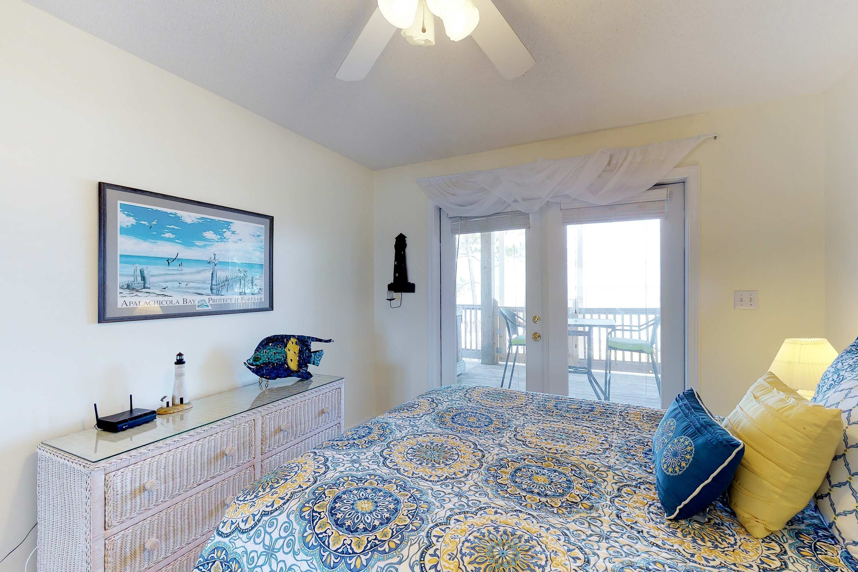 A bedroom with a patterned bedspread, a dresser with a decorative item, a framed picture on the wall, and a balcony with a chair.