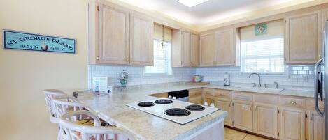 A kitchen with light wood cabinets, a white island with a stove, and a dining area with wicker chairs.