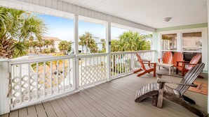 Een overdekte veranda met witte leuningen, houten stoelen en uitzicht op palmbomen en huizen.
