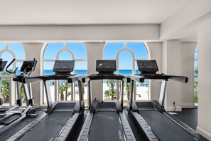 Un gimnasio con cintas de correr y equipo de ejercicio, ventanas amplias con vistas a la playa y palmeras.