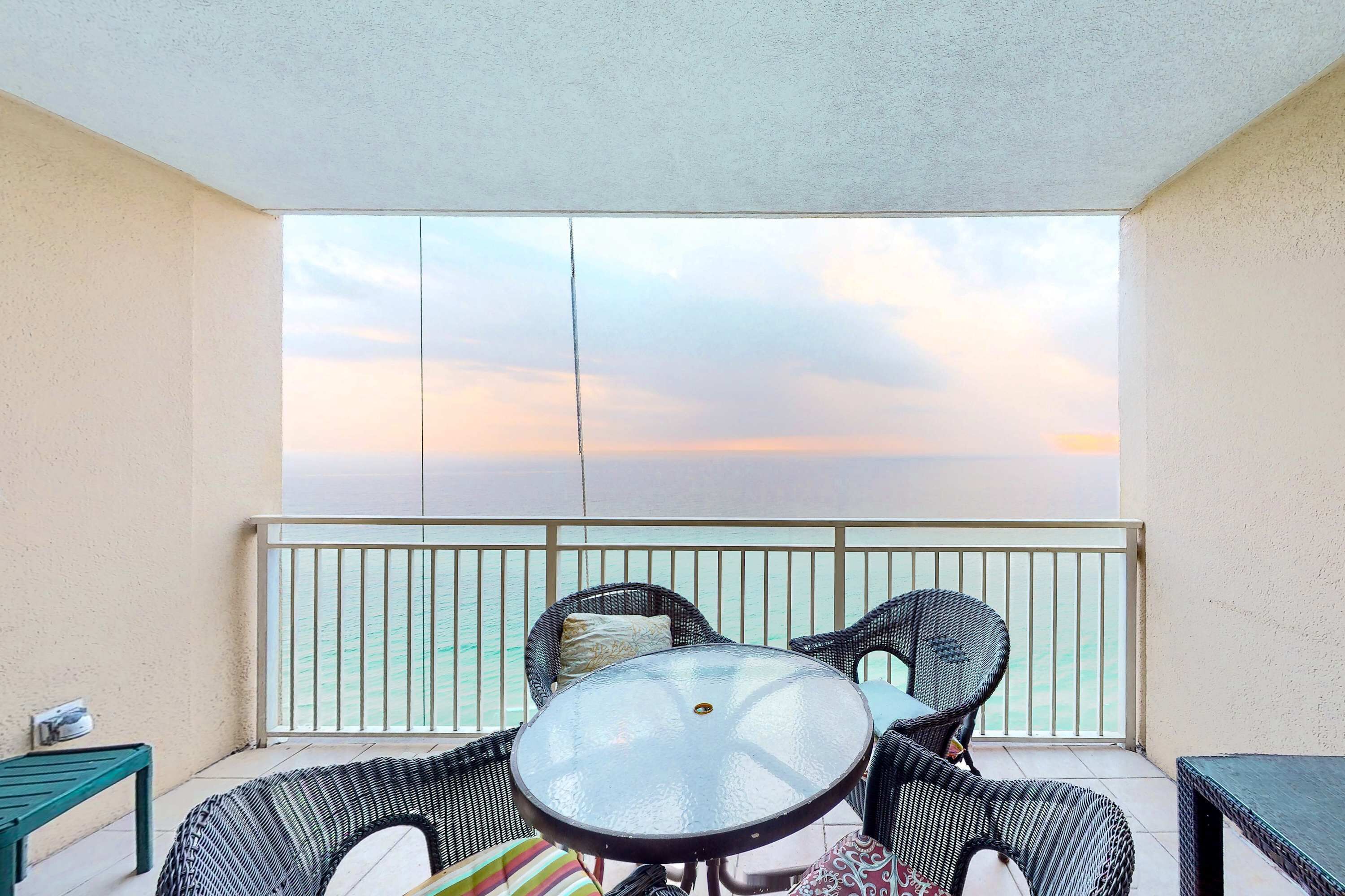 A balcony with wicker furniture, a glass table, and a view of the sea at sunset.