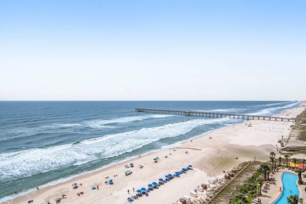 A beach with a long pier extending into the ocean, rows of umbrellas, and a swimming pool. - Beachfront resort condo with Gulf view, pools & gym - walk to Pier Park (Panama City Beach)