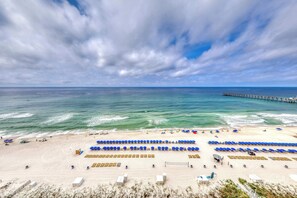 A beach with rows of blue umbrellas and sun loungers, a pier extending into the ocean, and a sandy beach area. - Beachfront resort condo with Gulf view, pools & gym - walk to Pier Park (Panama City Beach)