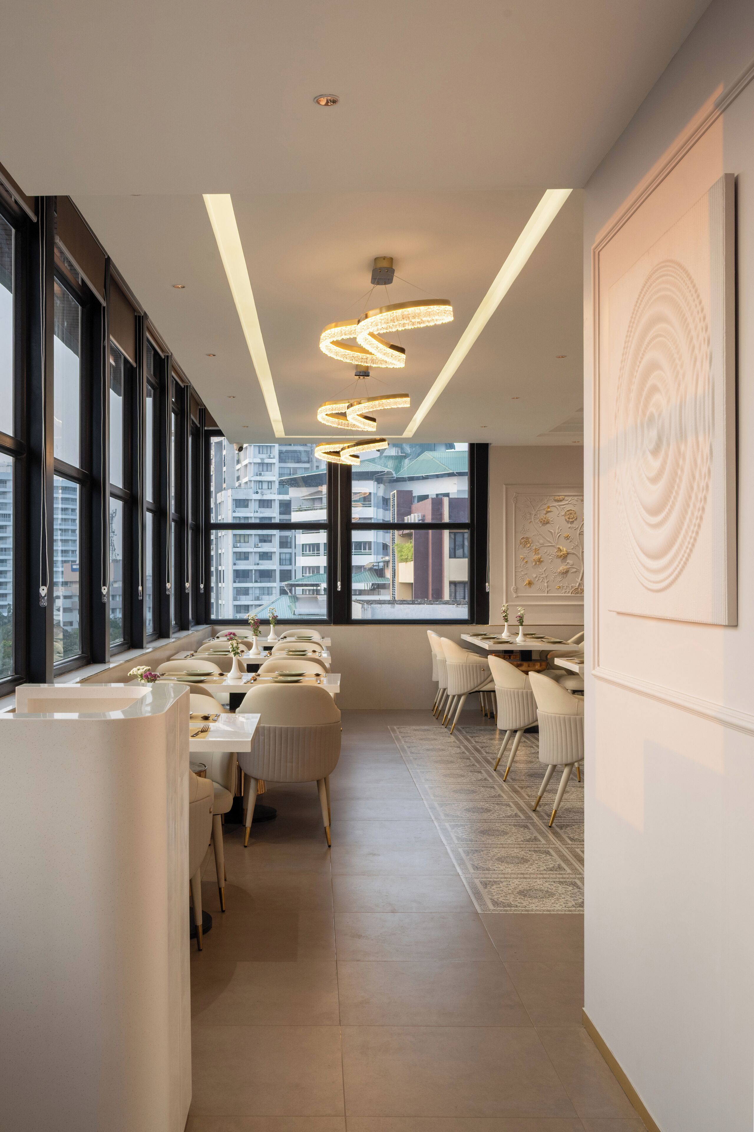 A modern dining area with long tables and chairs, large windows, and a decorative light fixture.