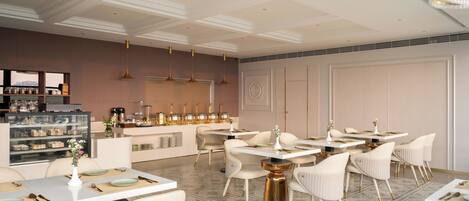 Un comedor moderno con decoración blanco y dorado, con varias mesas y sillas, una estación de servicio central y un piso estampado.