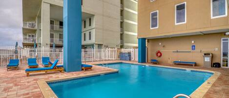 En swimmingpool med blÄ solstolar, ett vitt staket och en byggnad med flera balkonger i bakgrunden.